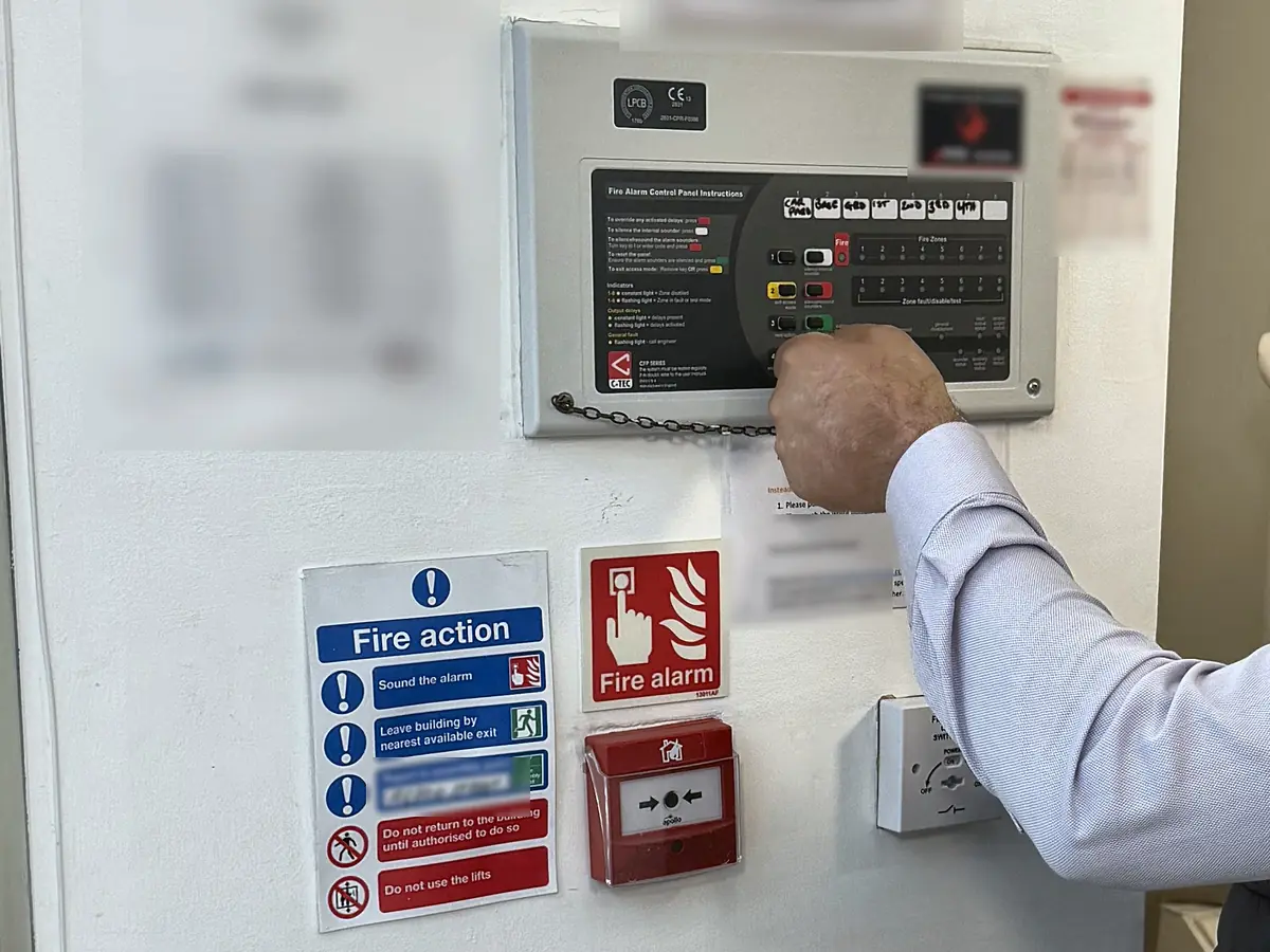 A conventional fire alarm panel tested on a service visit in the communal hallway of a London residential block, with the fire action notice and call point below