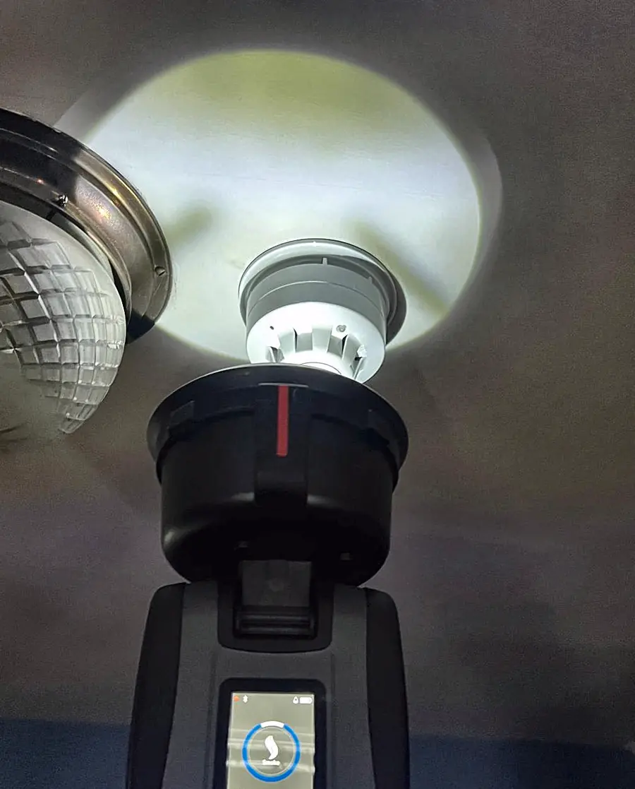 A smoke tester over a ceiling detector beside a cut-glass light, its screen showing the smoke test
