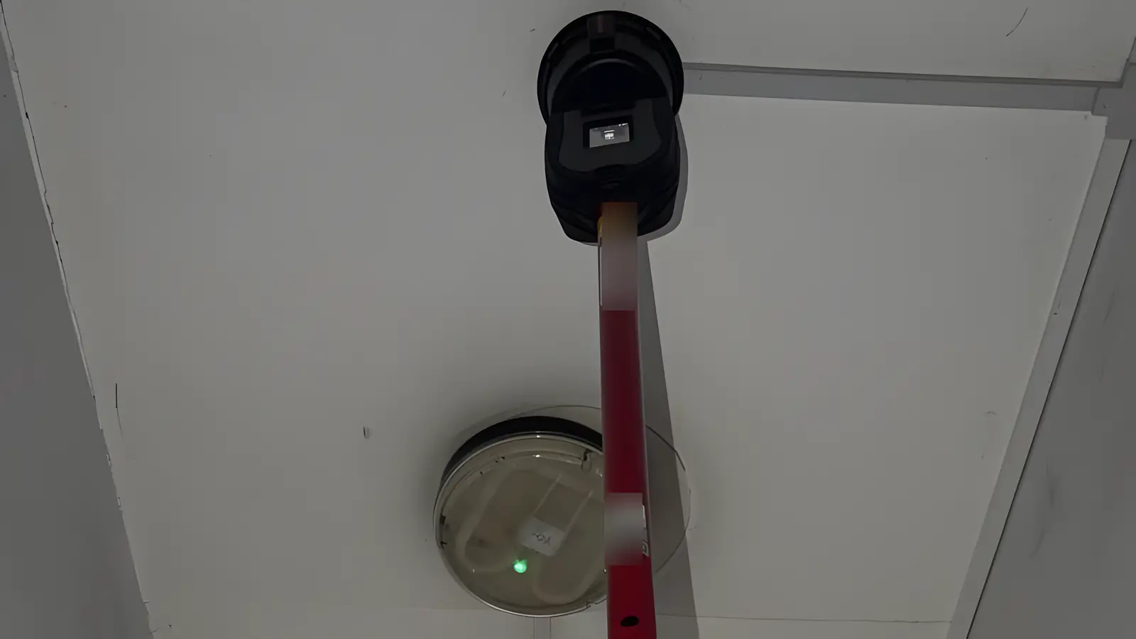 Fire alarm maintenance for managing agents in London: a smoke detector tested with a tester on a pole, under an emergency light in a corridor
